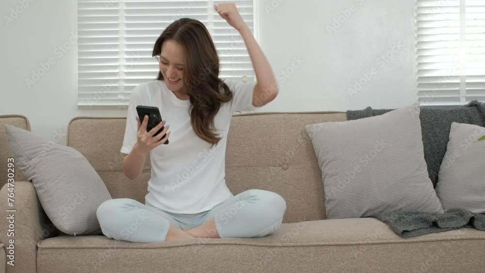 Young asian woman celebrating success holding mobile phone sitting on sofa in living room. Young asian woman celebrating success holding mobile phone sitting on sofa in living room.