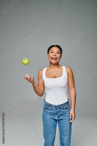 happy african american woman with white teeth throwing green apple in air on grey background