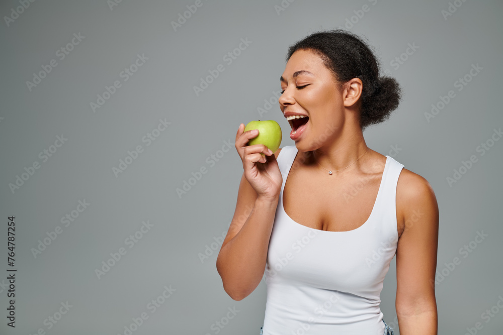 happy african american woman with white teeth biting green apple on grey background, healthy eating happy african american woman with white teeth biting green apple on grey background, healthy eating