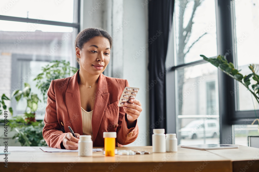 curly african american nutritionist evaluating medication while writing notes in her office curly african american nutritionist evaluating medication while writing notes in her office