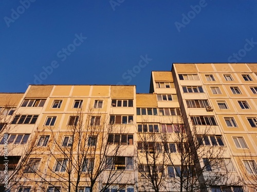 A bottom-up view of a multi-storey multi-apartment high yellow panel building with a large number of transparent windows, illuminated by sunlight, against which the sky is blue