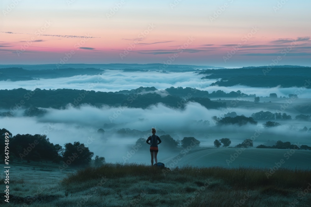 A runner pauses on hilltop, overlooking foggy valley below during early morning