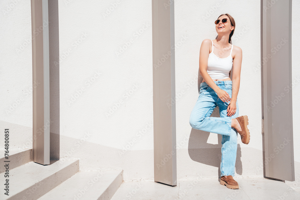 Beautiful smiling model in sunglasses. Female dressed in summer hipster white T-shirt and jeans. Posing near white wall in the street. Funny and positive woman having fun outdoors, in sunglasses Beautiful smiling model in sunglasses. Female dressed in summer hipster white T-shirt and jeans. Posing near white wall in the street. Funny and positive woman having fun outdoors, in sunglasses