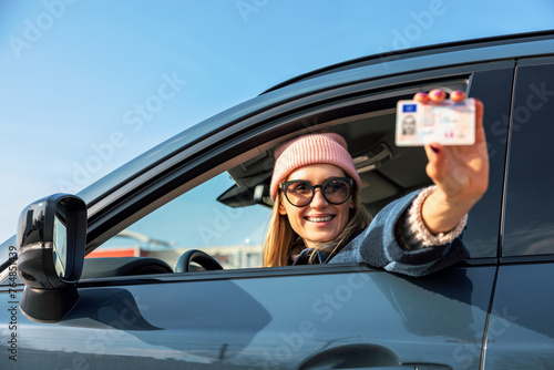 smiling woman showing her new driver license out of car window on sunny day