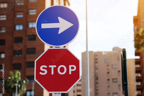 Traffic rules. Red road sign stop and blue and white right directional arrow on residential buildings background. Street sign direction pointing turn right, but need to stop before. Two road signs. © Andriy Medvediuk