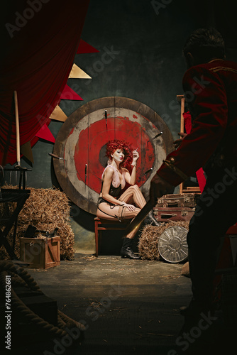Young redhead woman with makeup and costume sitting and smoking near target over dark retro circus backstage background. Man throwing knives. Concept of circus, performance, show, retro and vintage