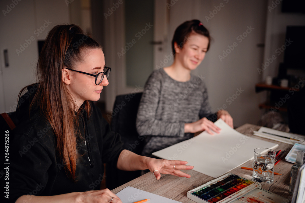 Two women share a creative moment; one chooses paper while the other prepares to paint, a symbol of inclusive artistic expression