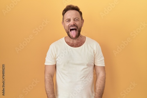 Middle age man with beard standing over yellow background sticking tongue out happy with funny expression. emotion concept.