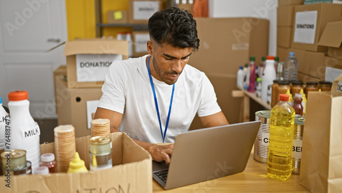 A focused hispanic man with a beard volunteers at a food donation center, managing inventory on a laptop.
