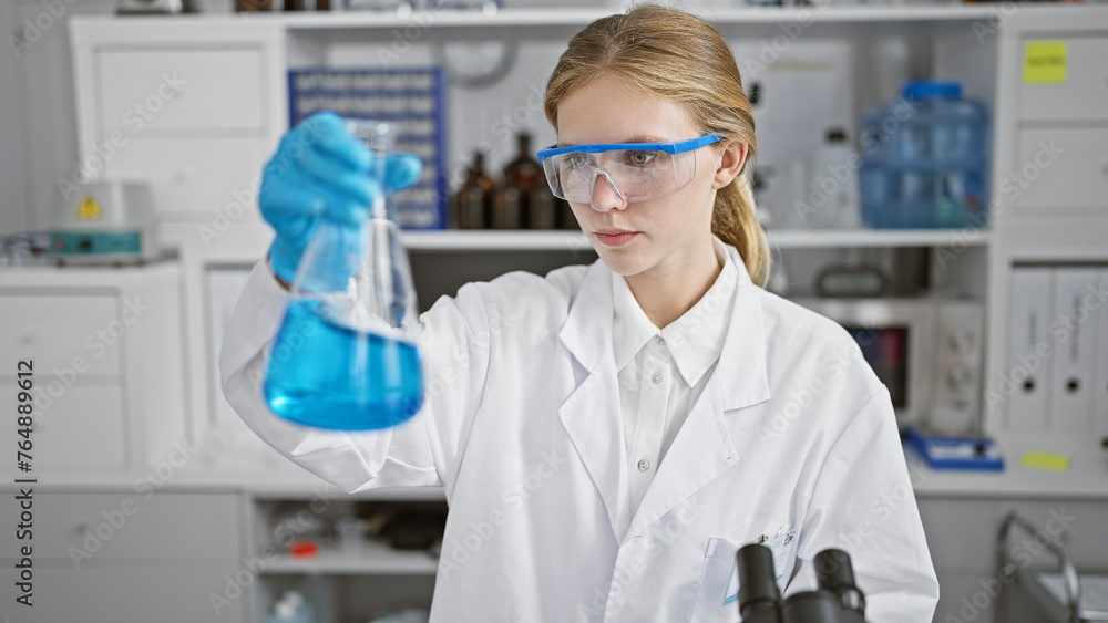 Fototapeta premium Blonde woman scientist examining a blue liquid in a flask in a laboratory setting