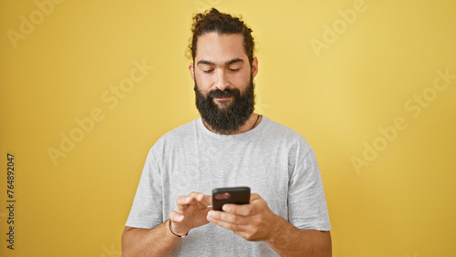 A young hispanic man with a beard smiles while using a smartphone against a yellow wall. © Krakenimages.com