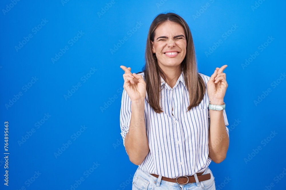 Hispanic young woman standing over blue background gesturing finger crossed smiling with hope and eyes closed. luck and superstitious concept. Hispanic young woman standing over blue background gesturing finger crossed smiling with hope and eyes closed. luck and superstitious concept.