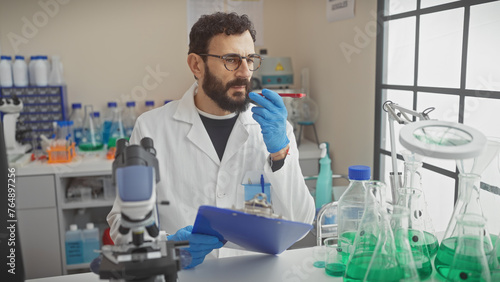 Middle-aged bearded man in lab coat examines a test tube inside a well-equipped laboratory with clipboard in hand. photo