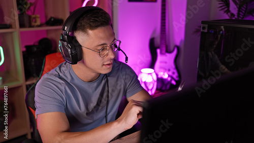 A focused young man using headphones works attentively in a dimly lit home office with gaming setup and decor. © Krakenimages.com