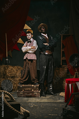 Retro-themed event. High African man standing with little servant boy over dark retro circus backstage background. Concept of circus, theater, performance, show, retro and vintage