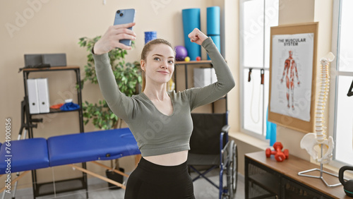 A young woman takes a selfie while flexing her arm in a modern rehab clinic's therapy room, exuding confidence and health.