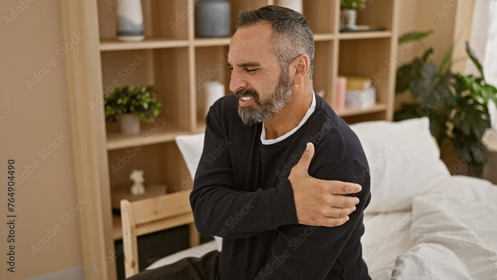 A mature hispanic man with grey beard in pain, holding his shoulder indoors, expressing discomfort in a bedroom setting. A mature hispanic man with grey beard in pain, holding his shoulder indoors, expressing discomfort in a bedroom setting.