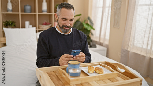Mature man with beard using smartphone in bedroom with breakfast tray.