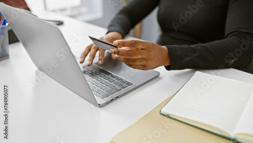 African american woman with credit card performing online transaction on laptop in modern office.