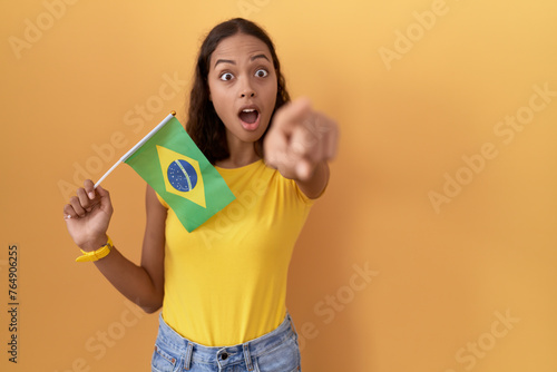 Young hispanic woman holding brazil flag pointing with finger surprised ahead  open mouth amazed expression  something on the front