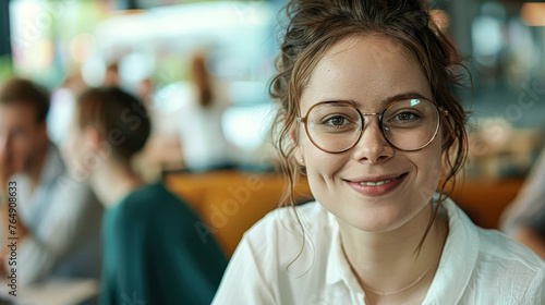 A woman wearing glasses is smiling at the camera. She is sitting at a table with other people