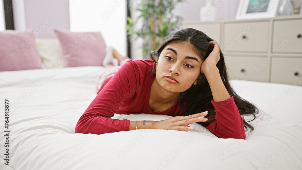 A thoughtful young indian woman with long hair lying on a bed, looking away in a well-lit bedroom. A thoughtful young indian woman with long hair lying on a bed, looking away in a well-lit bedroom.