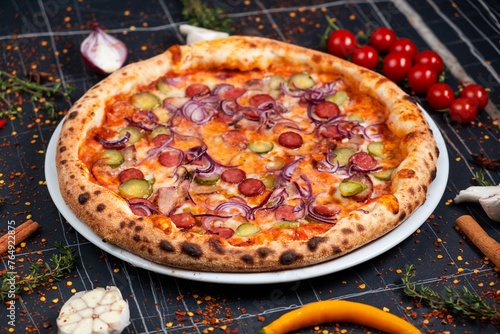 Oven baked pizza with mozzarella, ham, sausages, smoked chicken, red onion, tomatoes and pickled cucumbers over wooden background. Close up view