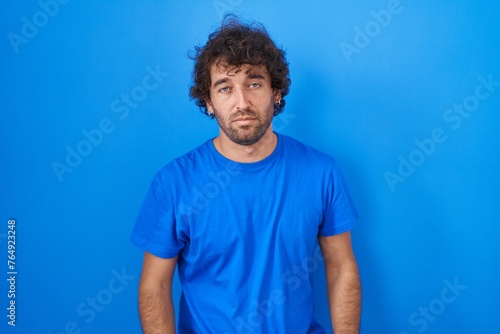 Hispanic young man standing over blue background looking sleepy and tired, exhausted for fatigue and hangover, lazy eyes in the morning. photo