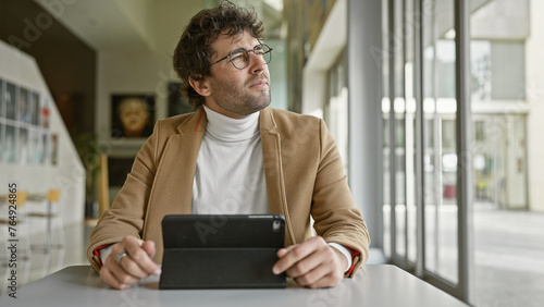 Thoughtful hispanic man with glasses in a modern office holding tablet