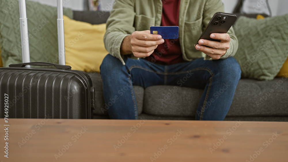 A young man sitting indoors with a suitcase, holding a credit card and smartphone, possibly preparing for travel or online shopping.