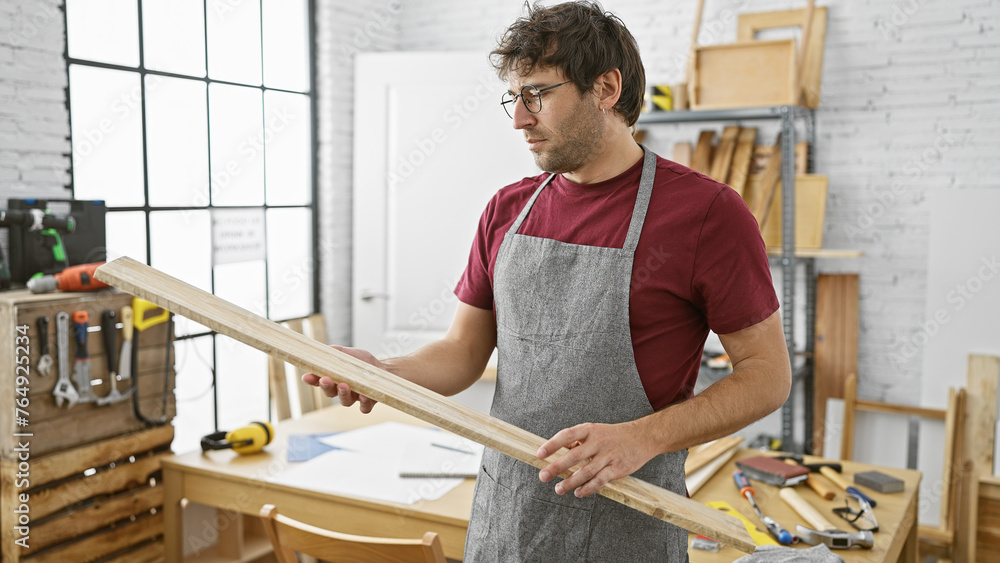 Hispanic man inspects wood in a sunny carpentry workshop wearing apron and glasses. Hispanic man inspects wood in a sunny carpentry workshop wearing apron and glasses.