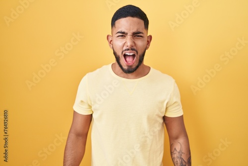 Young hispanic man standing over yellow background angry and mad screaming frustrated and furious, shouting with anger. rage and aggressive concept.