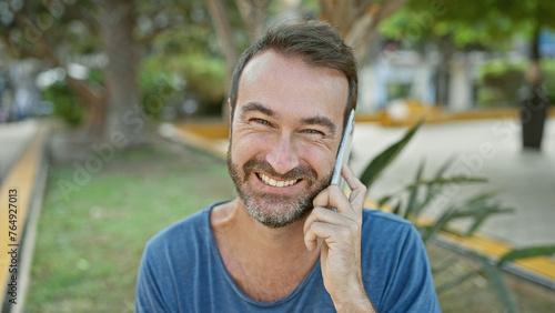 A cheerful middle-aged hispanic man with a beard making a phone call in a sunny park.