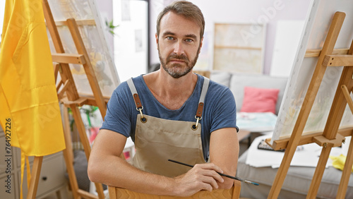 Portrait of a confident middle-aged man, artist, indoors, holding a paintbrush and wearing an apron.
