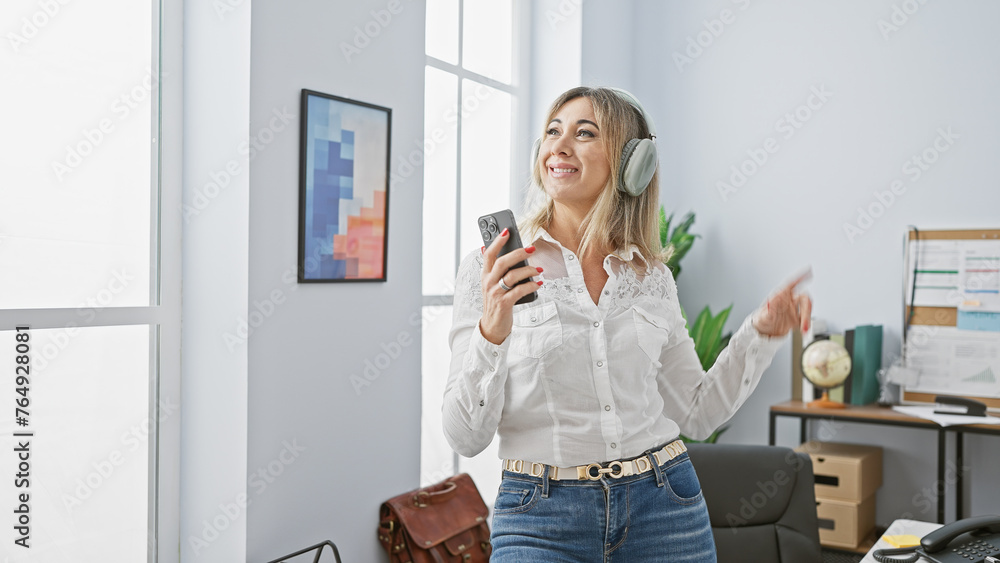 A joyful middle-aged woman enjoys music on headphones while holding a smartphone in a modern office. A joyful middle-aged woman enjoys music on headphones while holding a smartphone in a modern office.