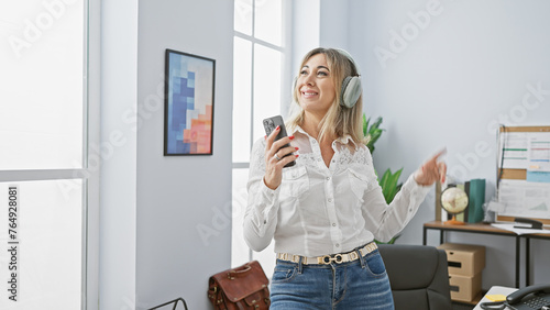 A joyful middle-aged woman enjoys music on headphones while holding a smartphone in a modern office.