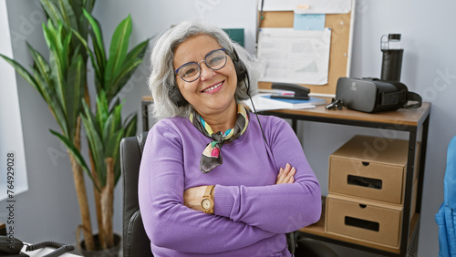 Fotografie Smiling mature woman with grey hair wearing headset in office, arms crossed, confident demeanor