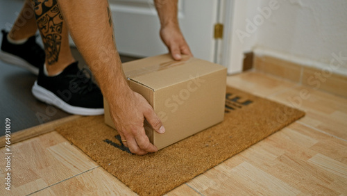 A tattooed man picks up a cardboard box from a doormat inside a home, signifying delivery or moving. © Krakenimages.com