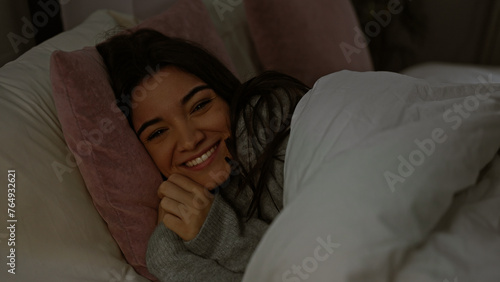Young hispanic woman smiling warmly, cuddled in bed at home during a cozy night.