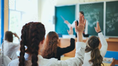 Intelligent group of young school children all raising their hands in the air to answer a question posed by the female teacher, view from behind