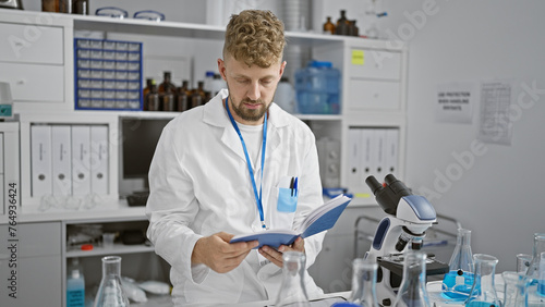 A young caucasian man with a beard reads a manual in a white laboratory coat indoors at a science lab