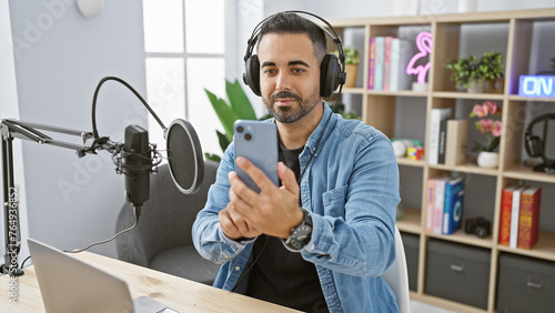Bearded hispanic man in studio with microphone and headphones holding smartphone, embodying podcasting or radio hosting.