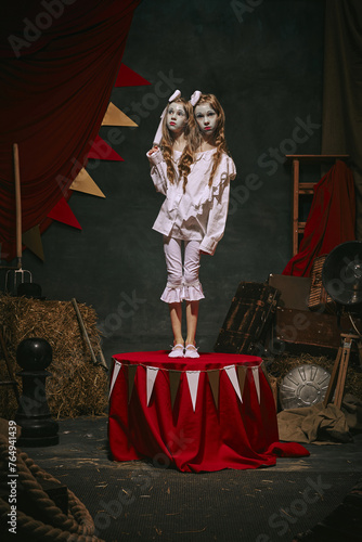 Siamese twin girls in white vintage costumes with makeup stand on stage on dark retro circus backstage background. Spooky-themed magic show. Concept of circus, theater, performance, retro and vintage