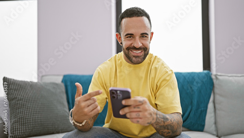 Smiling bearded man using smartphone in a cozy living room, conveying a casual and relaxed lifestyle.