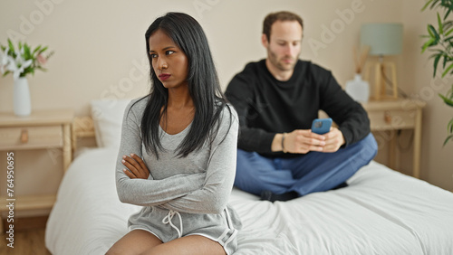 Beautiful couple sitting on bed with serious face while boyfriend using smartphone at bedroom