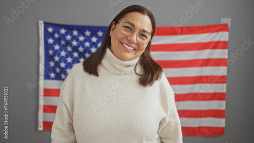 Smiling middle-aged hispanic woman in a white sweater standing before an american flag in an office setting photo