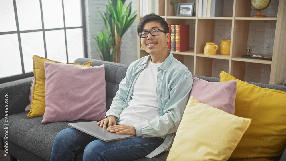 A young asian man wearing glasses, casually dressed, smiles while using a laptop on a gray couch in a well-lit living room with colorful pillows.