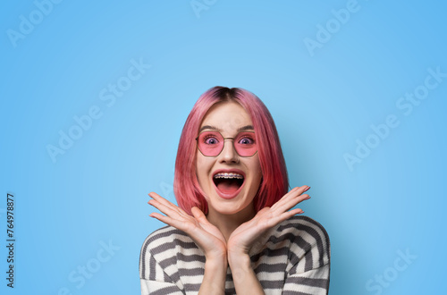 Excited surprised shock astonished very happy pink woman wear braces brackets sunglasses eye glass spectacles, open mouth, hold hands palms near face, isolated blue background. Mega sales ad concept