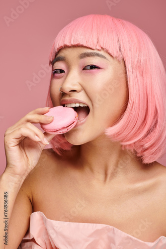 playful asian young woman with pink hair smiling and eating macaroon on vibrant background