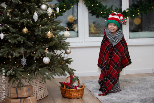 Happy little girl wearing a santa claus hat is wrapped in a warm checkered blanket in a christmas room interior.